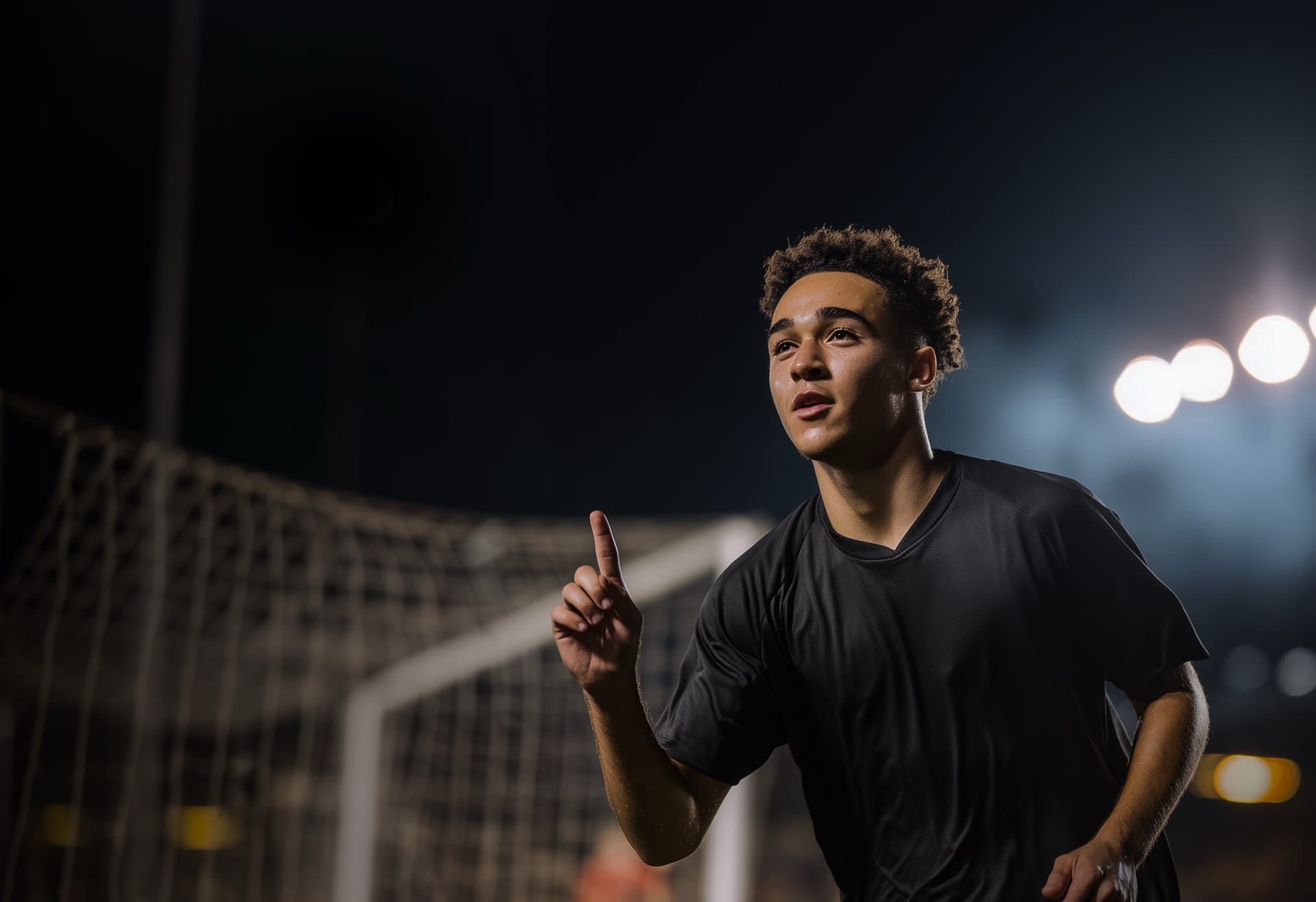 Young academy player pointing to the sky in front of a floodlit goal